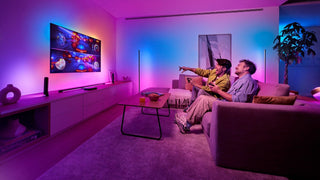 Twee mensen zitten op een bank in een woonkamer met kleurrijke sfeerverlichting en spelen opgewonden een videospel in twee delen op een tv aan de muur. De kamer is modern met een gezellige inrichting.