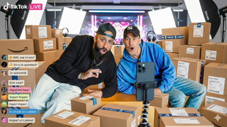 Twee opgewonden mannen die livestreamen op TikTok zitten tussen veel kartonnen dozen en reageren verbaasd. Reacties verschijnen in het Nederlands op het scherm terwijl ze filmen met een smartphone op een statief.