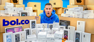 Een man met een blauwe capuchon lacht terwijl hij wordt omringd door dozen en verschillende elektronica van merken als Apple, Sony, JBL, Logitech en Xiaomi, waaronder telefoons, koptelefoons en opladers.