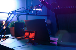 Een schemerig verlichte podcast- of radiostudio met een laptop, microfoon en een gloeiend rood ON AIR-teken op het bureau, omringd door moderne apparatuur en paarsblauwe verlichting.
