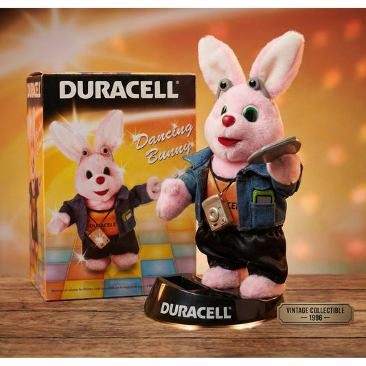 Een pluche Duracell Dancing Bunny uit 1996 (New Rare Old Stock) staat op een gemerkte voet, houdt een batterij vast en draagt een camera en spijkerjasje. De doos met het bijpassende konijntje staat op de achtergrond en is gemarkeerd als vintage collectible.